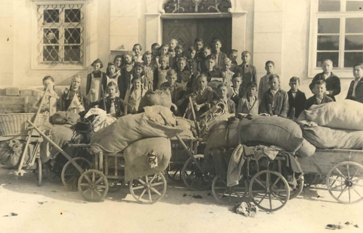 Historische Klassenaufnahme einer Schulklasse vor dem Rathaus in Straßberg mit Leiterwagen und gefüllten Säcken mit Stoffballen um das Jahr 1930